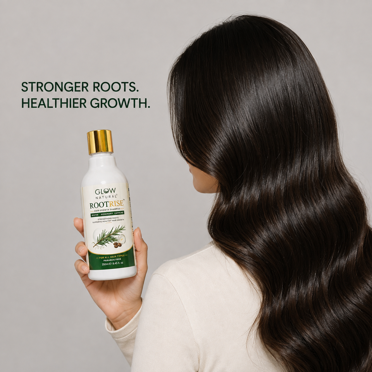 A woman with long, dark, glossy hair is shown from behind holding a bottle of Glow Naturally Rootline Hair Growth Serum. The background is a solid light gray, with promotional text on the left reading 'STRONGER ROOTS.